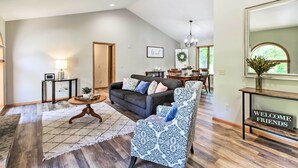 A spacious living room with a dark grey sofa, patterned armchair, and a rug. A 'WELCOME FRIENDS' sign is on a console table.