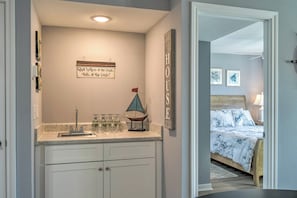 A bathroom with a sink, mirror, and towel rack. There is a bed with a blue and white patterned bedspread in the adjacent room.