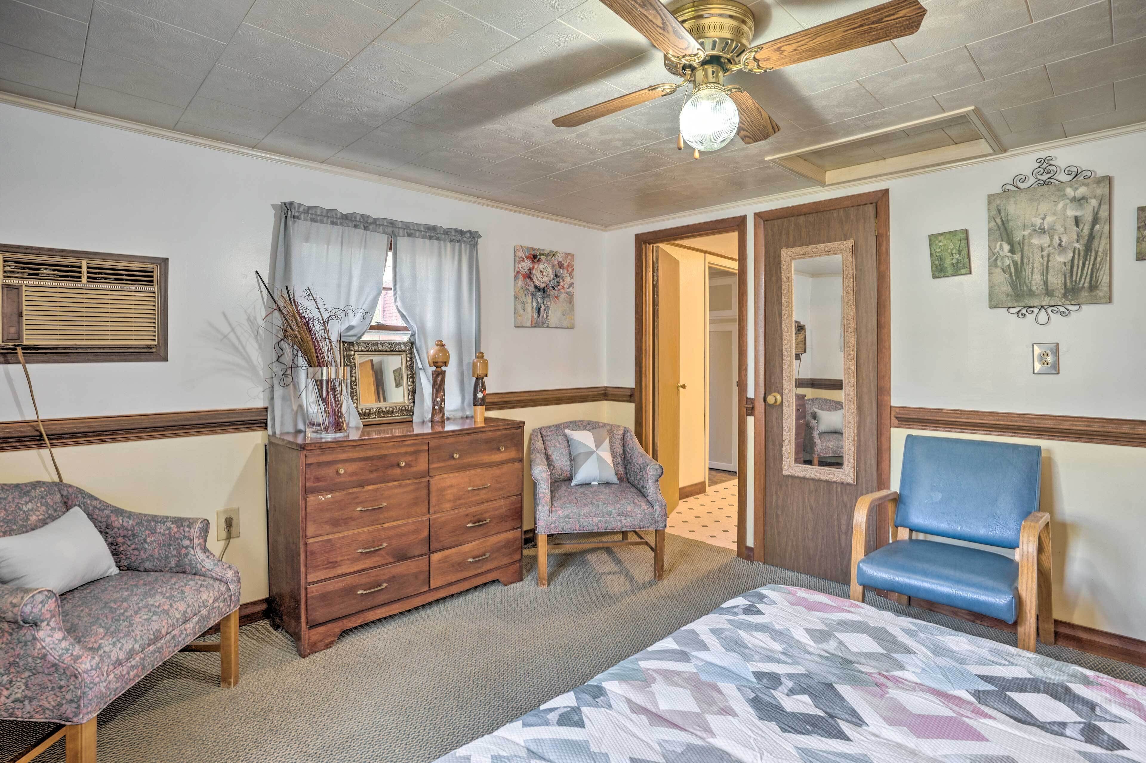 A bedroom with a bed, dresser, two chairs, a ceiling fan, and artwork on the walls.