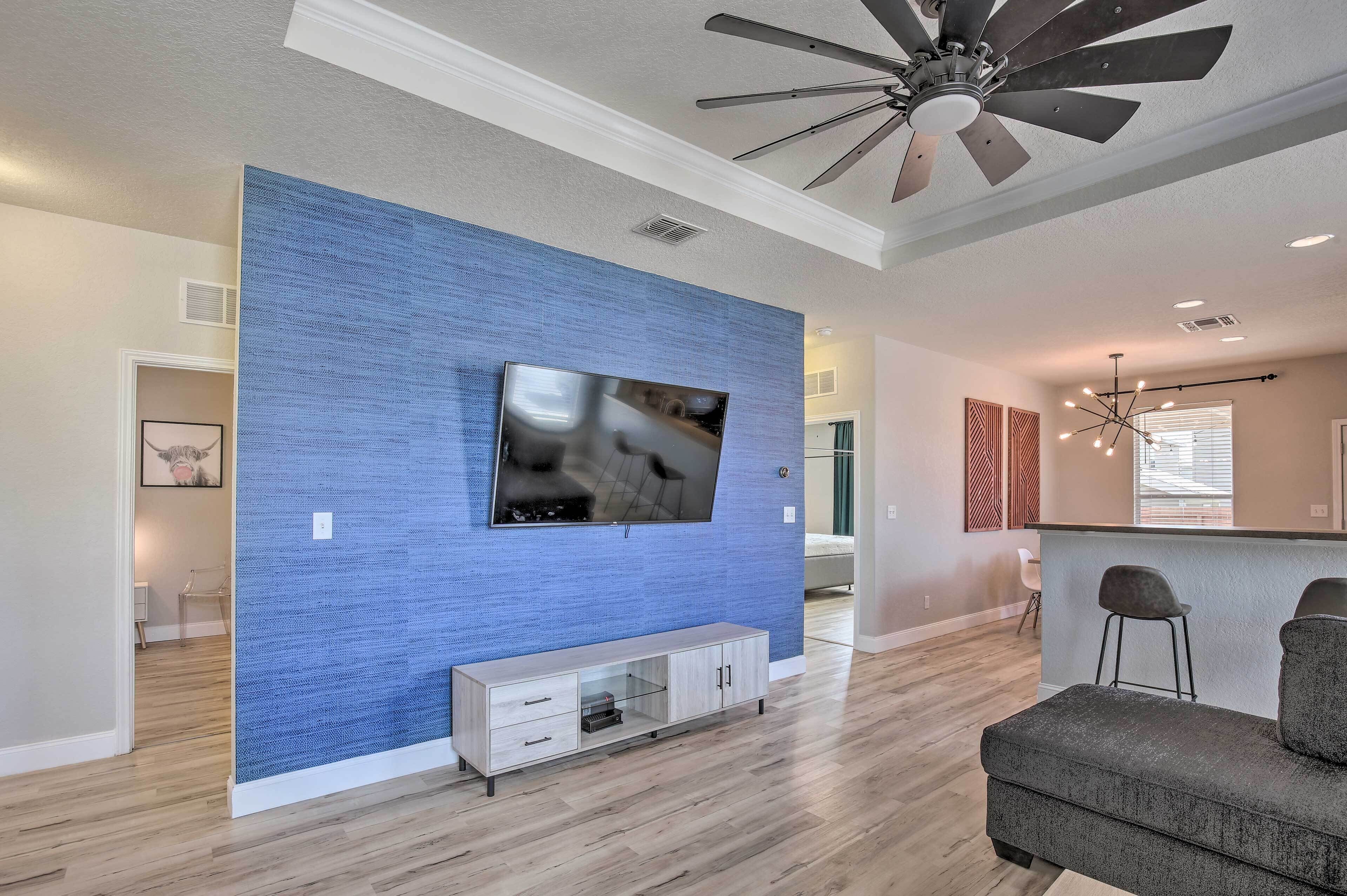 A modern living room with a flat-screen TV mounted on a textured blue wall, a wooden entertainment unit, a ceiling fan, and a grey sofa.
