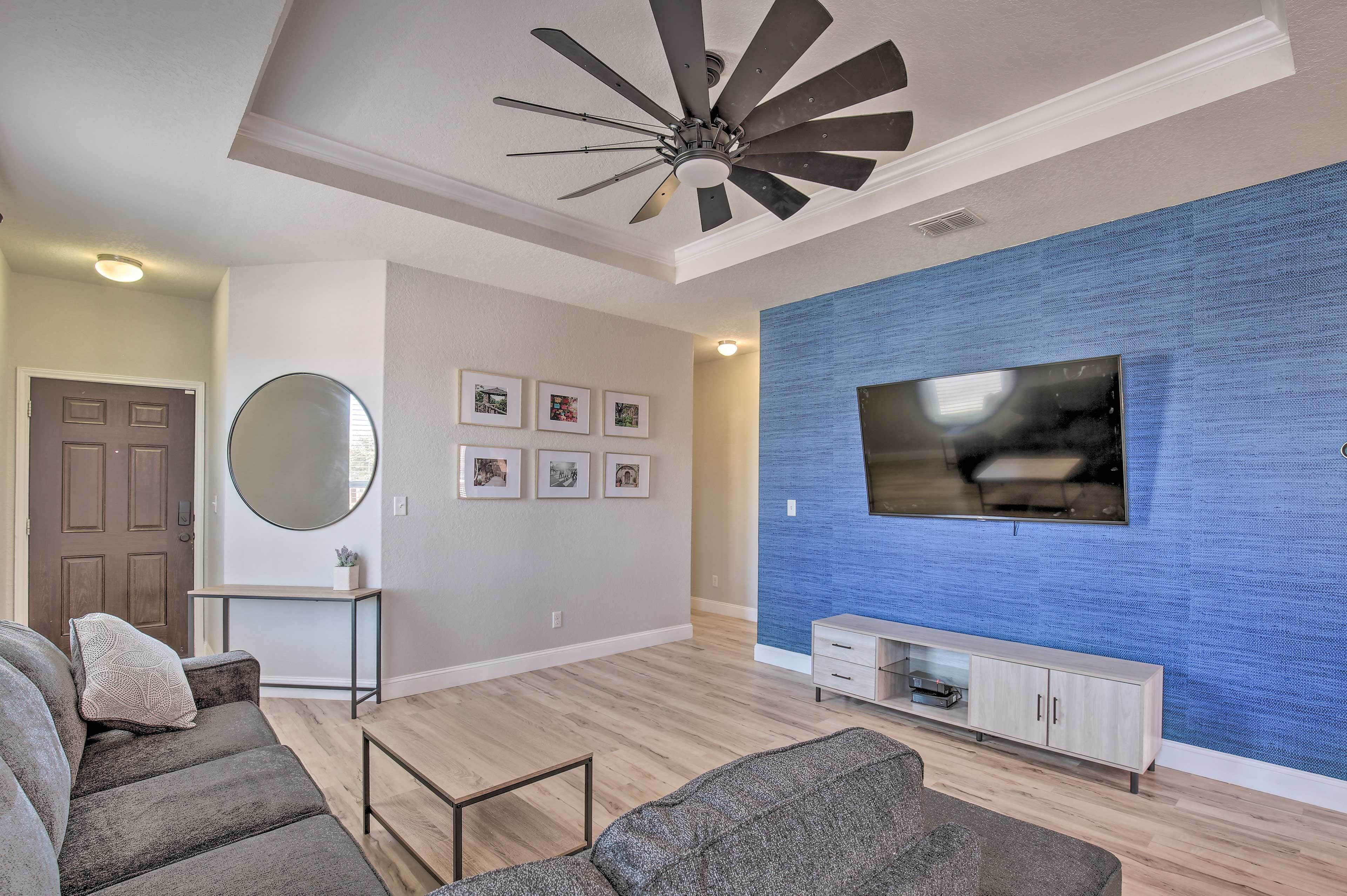 Una sala moderna con ventilador de techo, un televisor plano grande montado en una pared azul con textura, piso de madera y un sofá gris.
