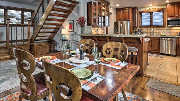 Un comedor con una mesa de madera dispuesta para cuatro personas, rodeada de sillas de madera. Se ve una escalera a la izquierda, que conduce a otro nivel. Se distingue la cocina al fondo, con armarios de madera y electrodomésticos de acero inoxidable.