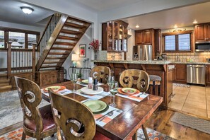 A dining area with a wooden table set for four, surrounded by wooden chairs. A staircase is visible to the left, leading to another level. The kitchen area is visible in the background with wooden cabinets and stainless steel appliances. - Downtown Park City Vacation Rental, Near Ski Lift! (Park City)