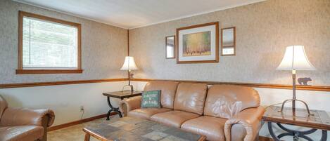 A living room with a leather sofa, a wooden coffee table, and two lamps. There is a window with blinds, a painting on the wall, and a mirror above the sofa.