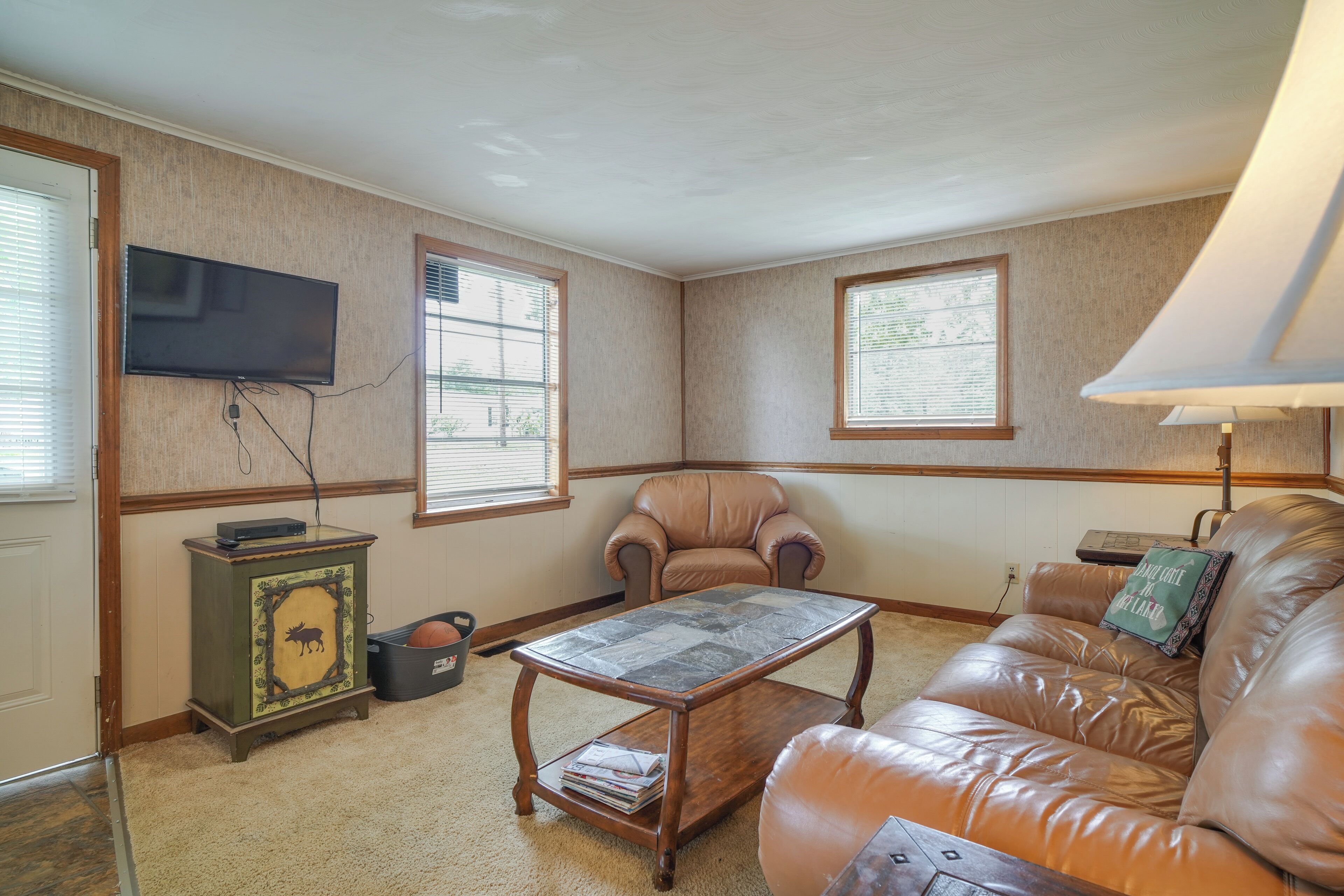 A living room with a leather couch, a coffee table, a television mounted on the wall, and a window with blinds.