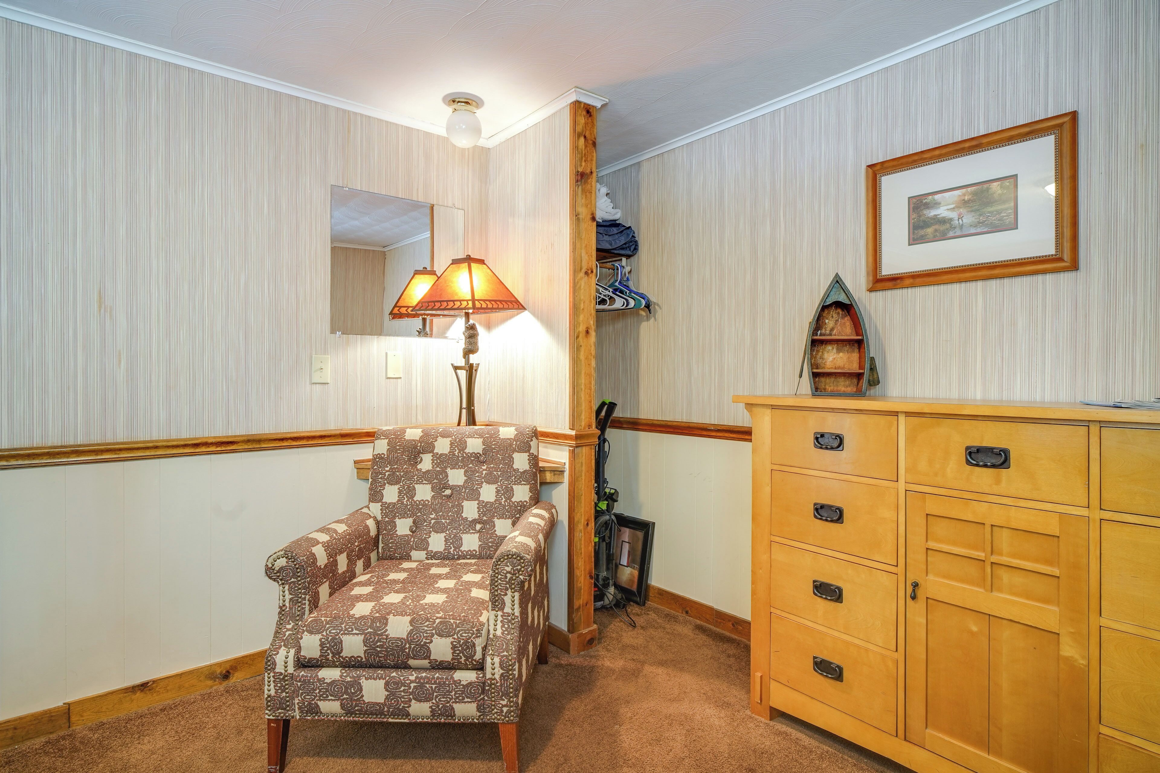 A room with a patterned armchair, a wooden cabinet with drawers, a lamp, a framed picture, and a small shelf with folded items.
