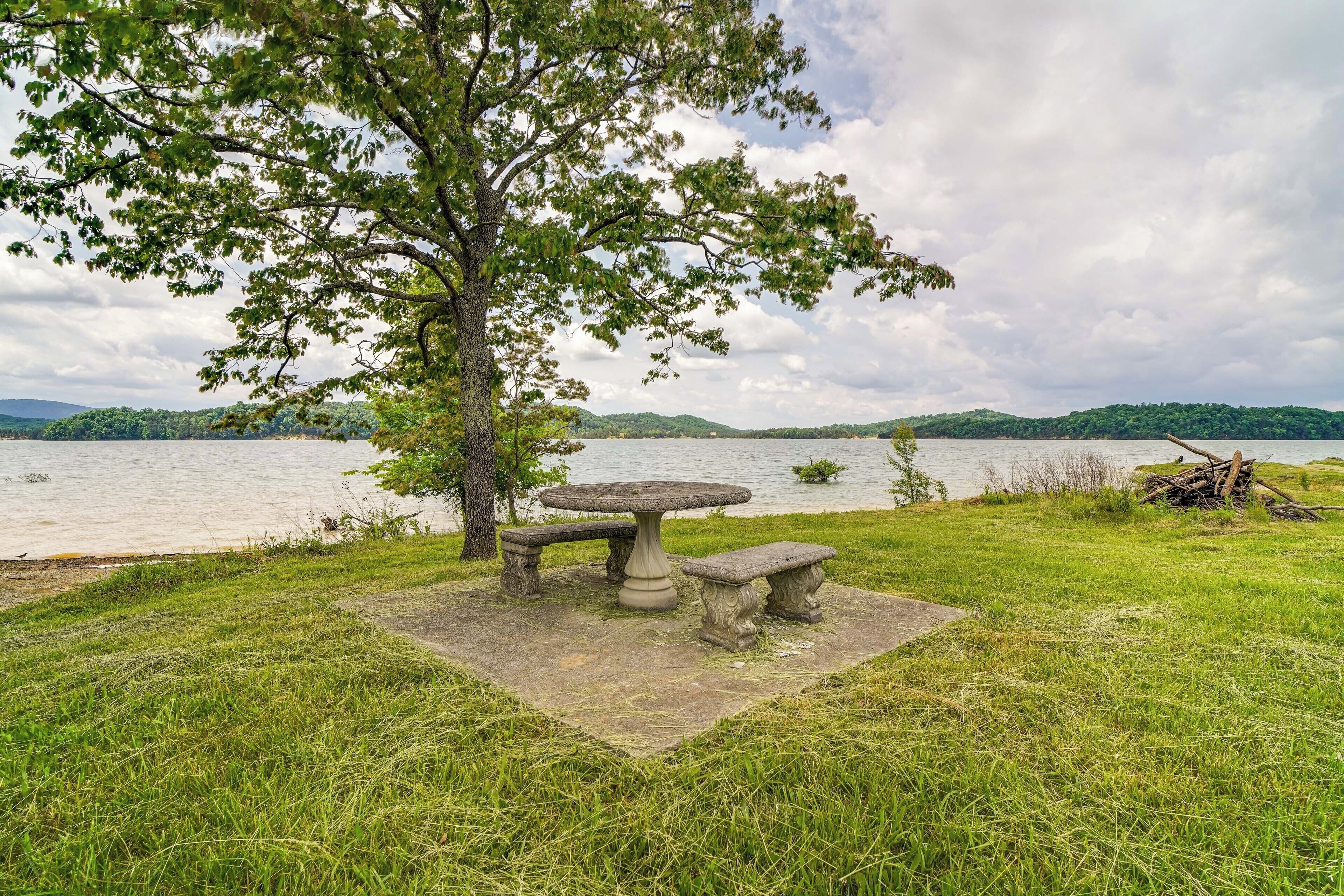 A stone table and benches set on a grassy area near a lake, with trees and hills in the background.