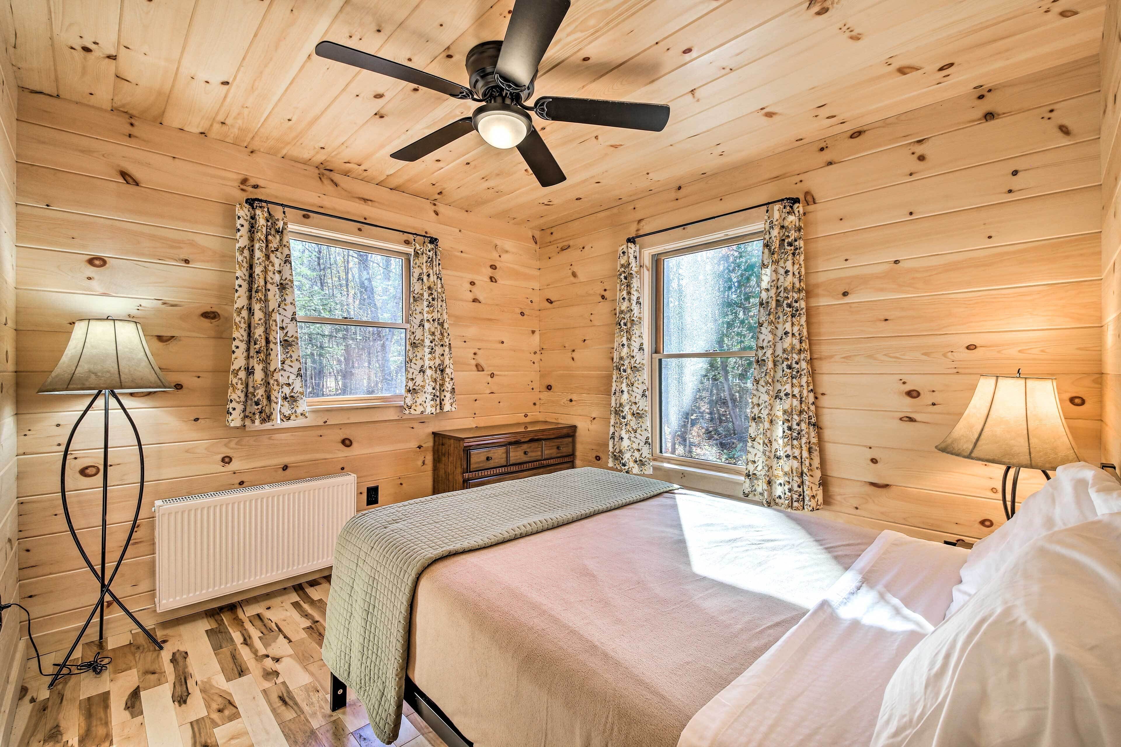 Une chambre à coucher dans un chalet en bois, avec un lit, deux fenêtres avec des rideaux, un ventilateur de plafond et une lampe de chevet.