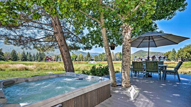 Un bain à remous sur une terrasse en bois, avec une table et des chaises, entouré d’arbres et offrant une vue sur une maison et des montagnes.