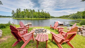 Ein Sitzbereich am Seeufer mit roten Adirondack-Stühlen um eine Feuerstelle, mit Blick auf einen ruhigen See mit einem Steg, umgeben von Bäumen.