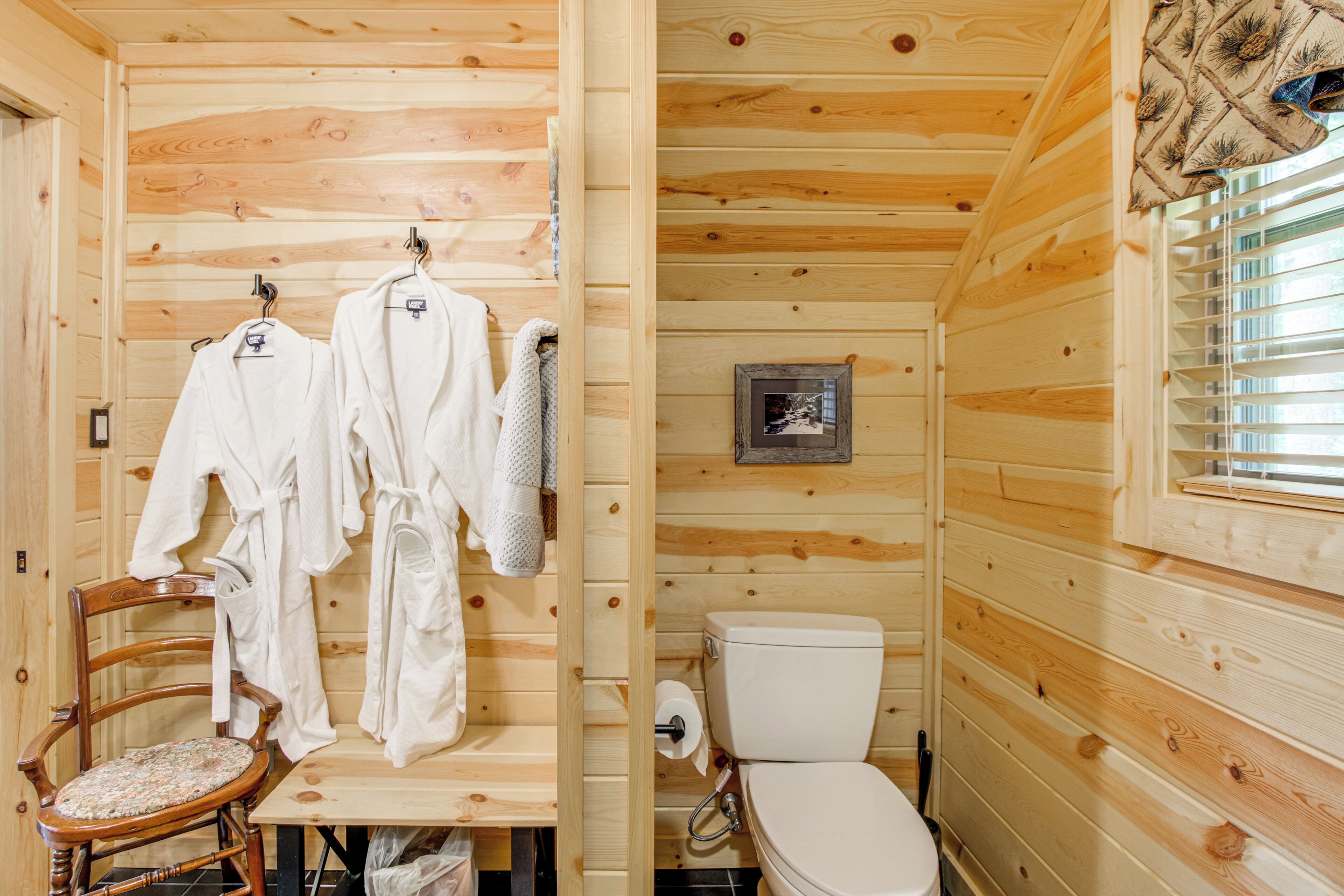 A wooden cabin bathroom with a toilet, a wooden chair, and a rack holding white robes.