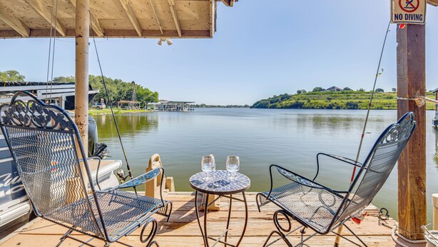Une terrasse en bois avec des chaises et une table en métal, donnant sur un lac paisible où un bateau est amarré à proximité.