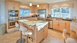 A modern kitchen with wooden cabinets, stainless steel appliances, a marble countertop, and a large island with bar stools. - Luxe Oceanfront Home w/ Cadillac Mountain Views! (Sullivan)
