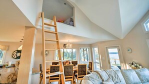 A spacious room with a loft bed, a dining area with a wooden table and chairs, and a view of the outdoors through large windows. - Luxe Oceanfront Home w/ Cadillac Mountain Views! (Sullivan)