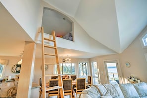 A loft bedroom with a high ceiling, a wooden ladder, a dining area with a table and chairs, and a view of the outdoors through large windows. - Luxe Oceanfront Home w/ Cadillac Mountain Views! (Sullivan)