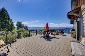 A wooden deck with outdoor furniture, a red umbrella, and a scenic view of mountains and water.
