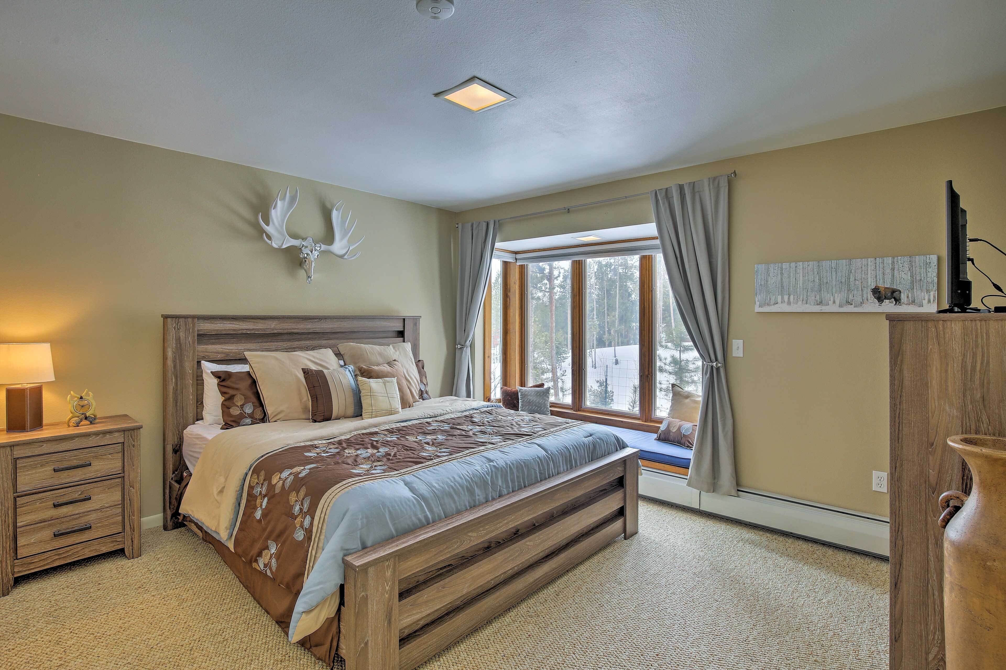A bedroom with a large bed, wooden headboard, a nightstand with drawers, a bedside table with a lamp, a window with curtains, and a wall-mounted deer head decoration.