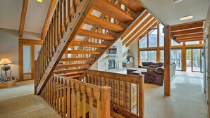 A spacious living area with a wooden staircase, a stone fireplace, and large windows offering views of the outdoors.