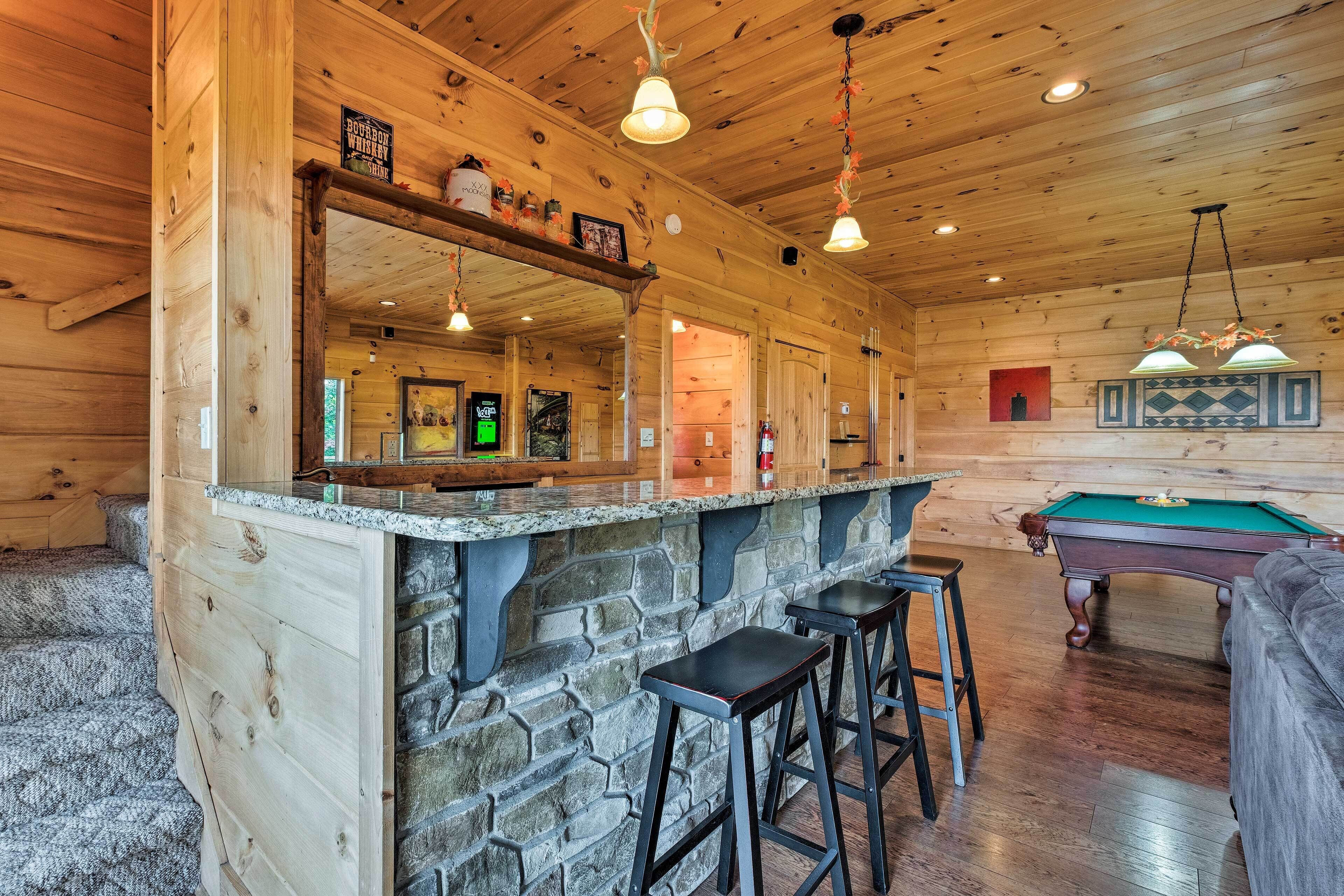 A wooden cabin interior with a bar area, stone counter, pool table, and bar stools.
