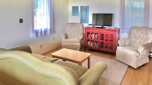 Un salon avec deux fauteuils beiges, une table basse en bois, une bibliothèque rouge et un téléviseur posé sur un meuble.