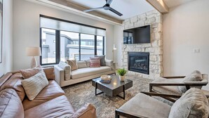 A modern living room with a stone fireplace, a ceiling fan, and a large window offering a view of the outside. - Mountain-view Townhome Near Powder Mountain! (Huntsville)