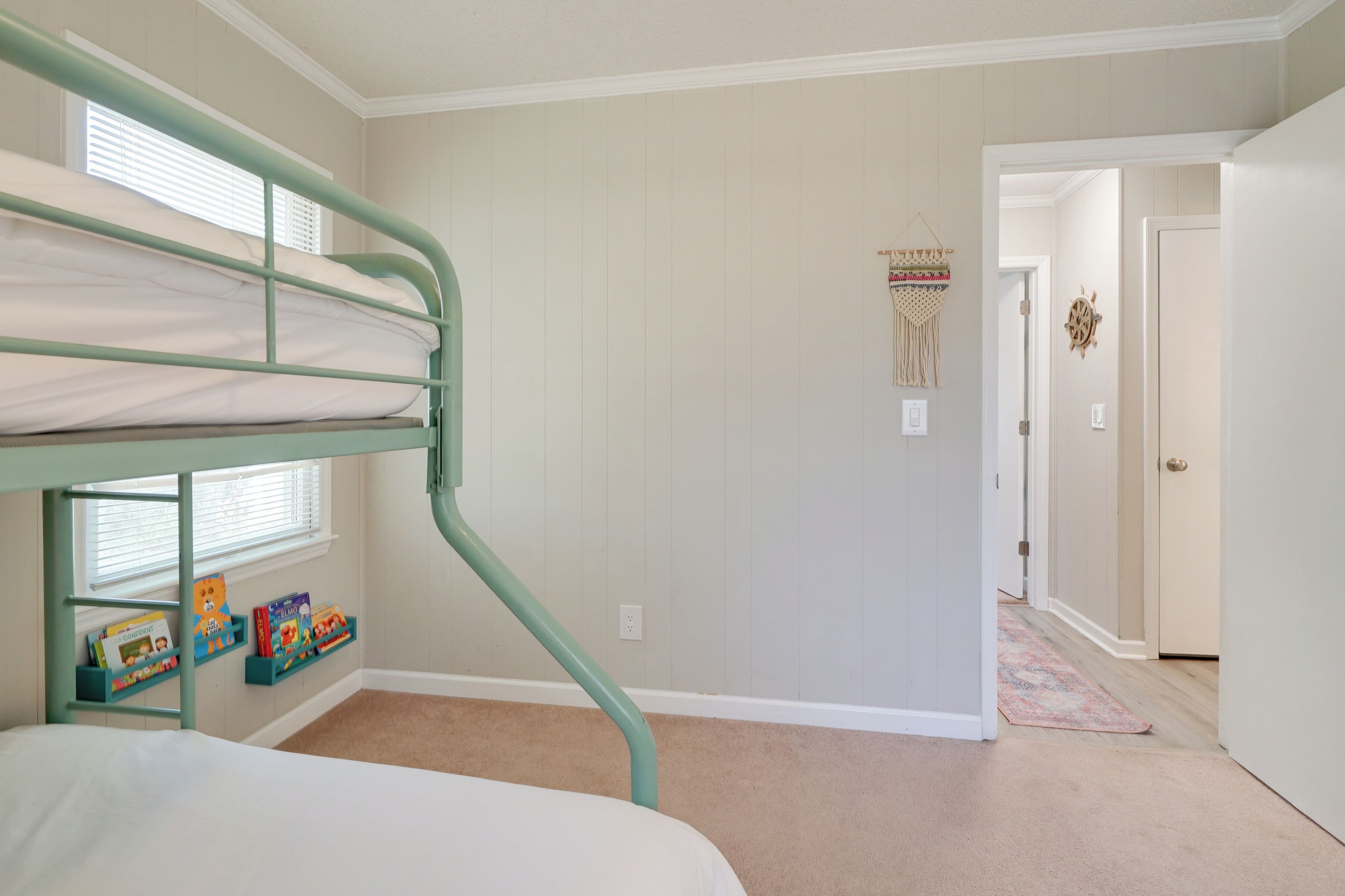 Una cama litera con estructura verde, una ventana con persianas y un estante fijado a la pared con libros infantiles.