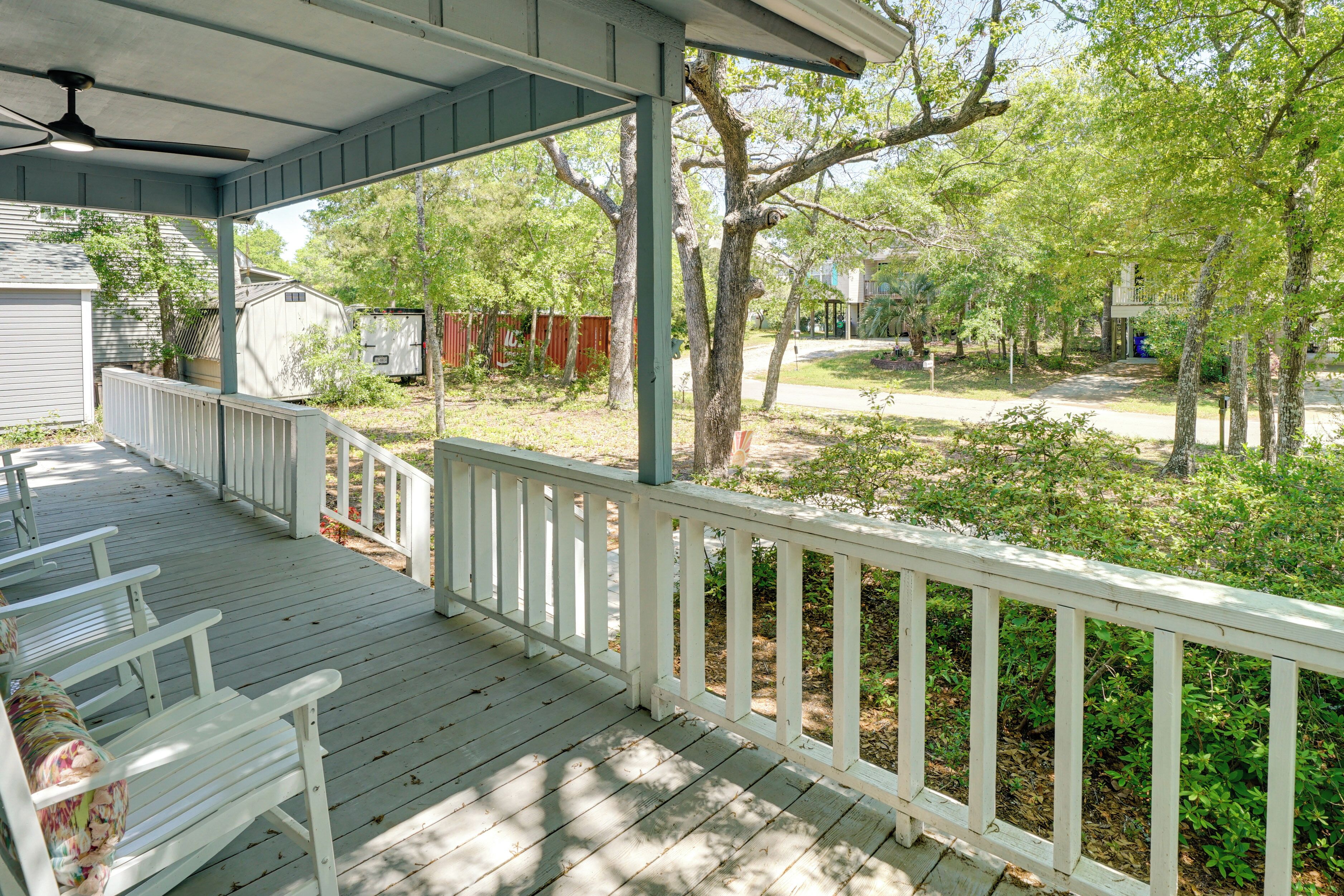 A covered porch with white railings, a ceiling fan, and a view of a residential area with trees and a fence.