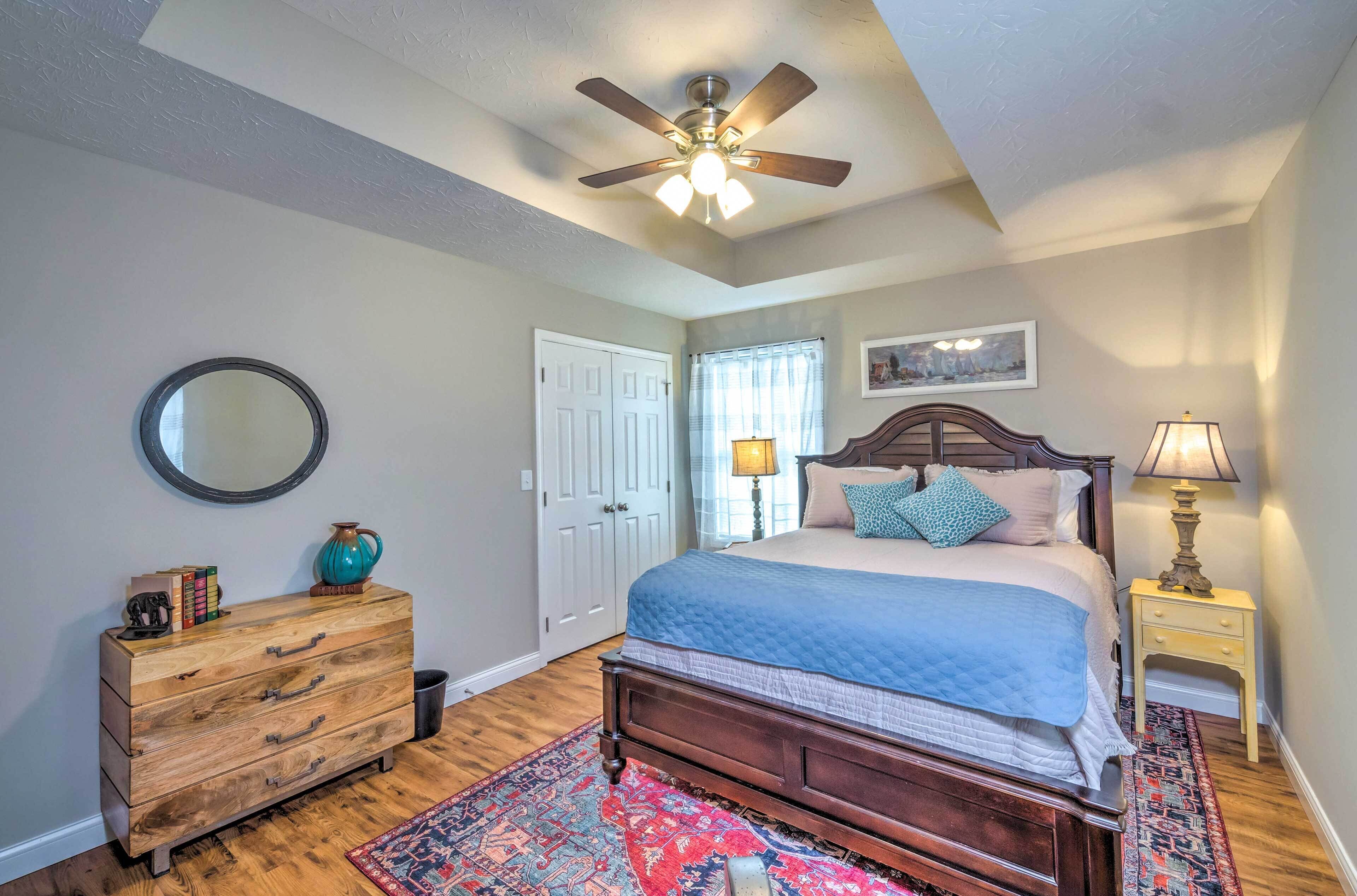 A bedroom with a wooden dresser, a bed with a blue quilt, a ceiling fan, a nightstand with a lamp, and a patterned rug.