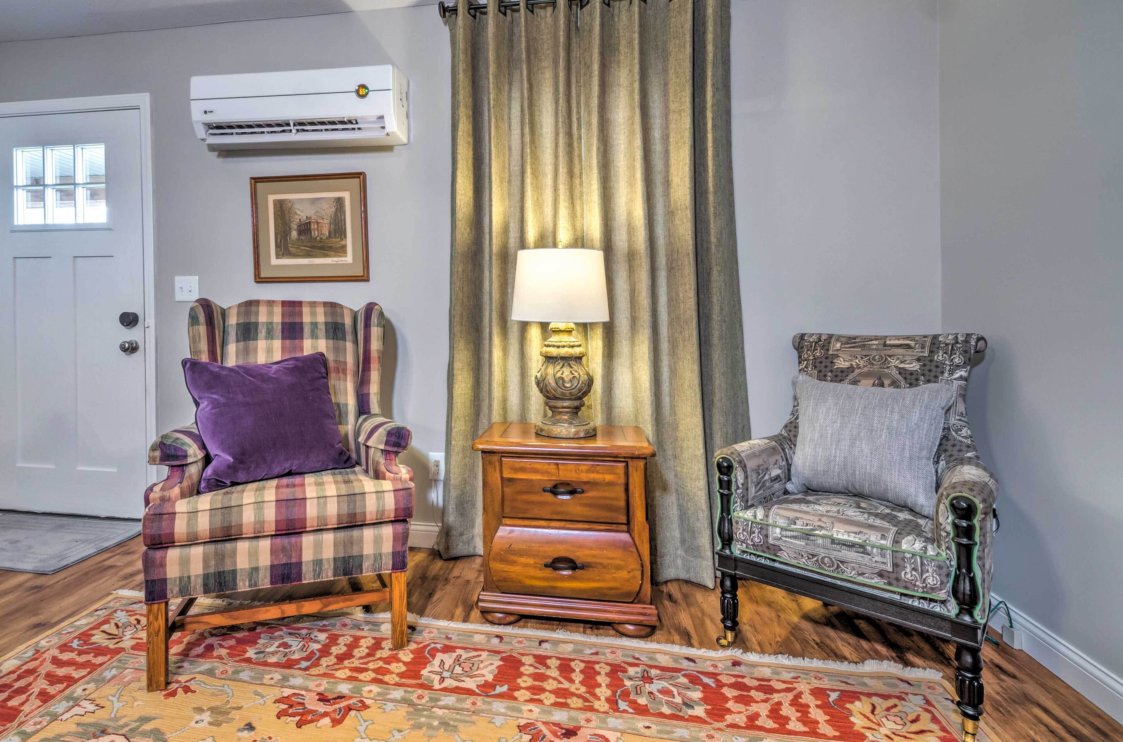 A room with a plaid armchair, a patterned armchair, a wooden nightstand with a lamp, and a red patterned rug.
