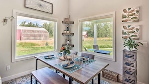Een eetgedeelte met een houten tafel voor vier personen, grote ramen en uitzicht op een schuur en groen buiten.