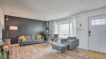 Un salon moderne avec un canapé gris, un pouf et un fauteuil, un plancher en bois et une grande fenêtre avec des rideaux.