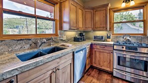A kitchen with wooden cabinets, a granite countertop, a dishwasher, a stove, and a window with a view of a house and trees. - Quiet Park City Home w/ Hot Tub - Walk to Main St! (Park City)