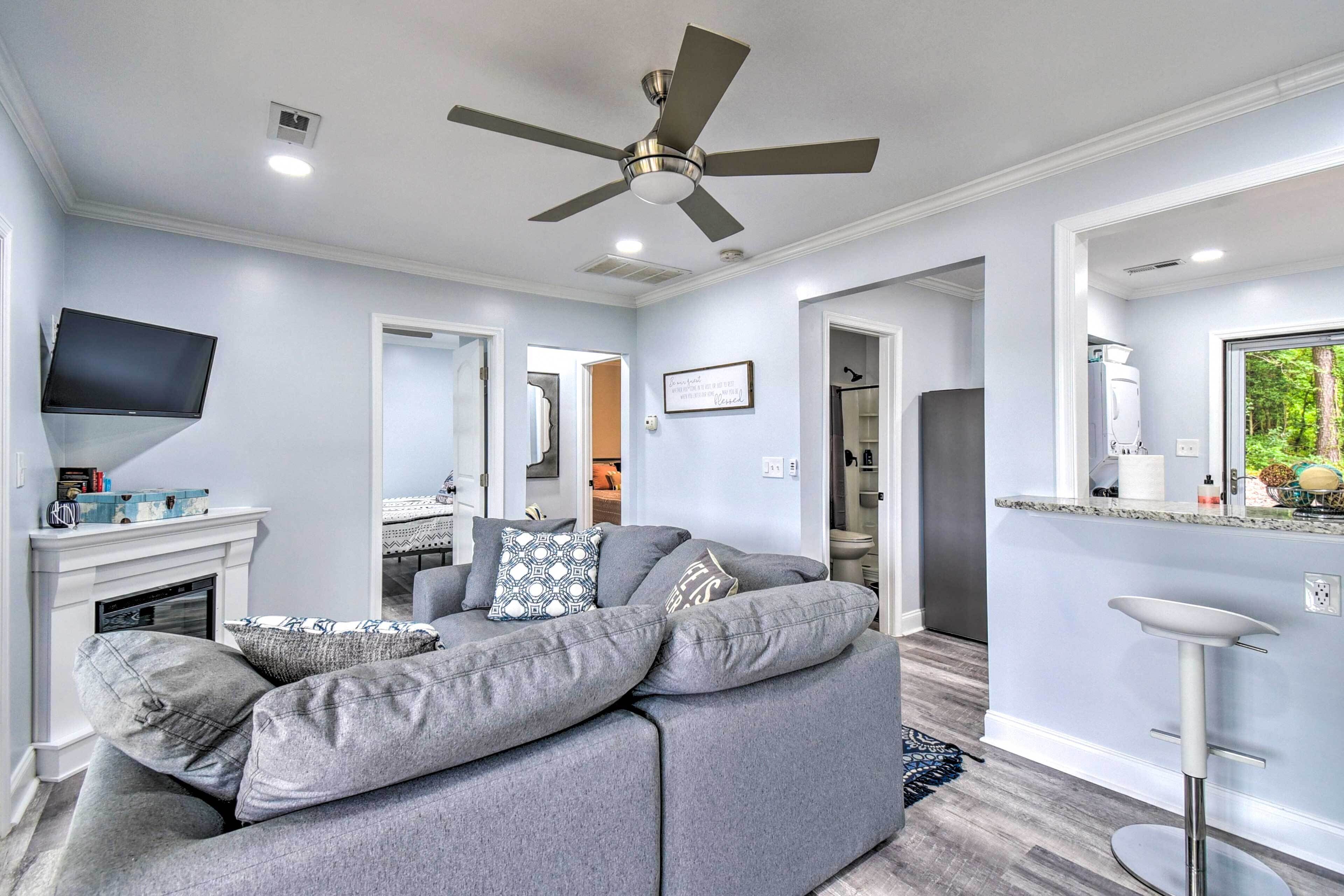 A modern living room with a grey sofa, a flat-screen TV, a ceiling fan, and a kitchen area with a bar stool.