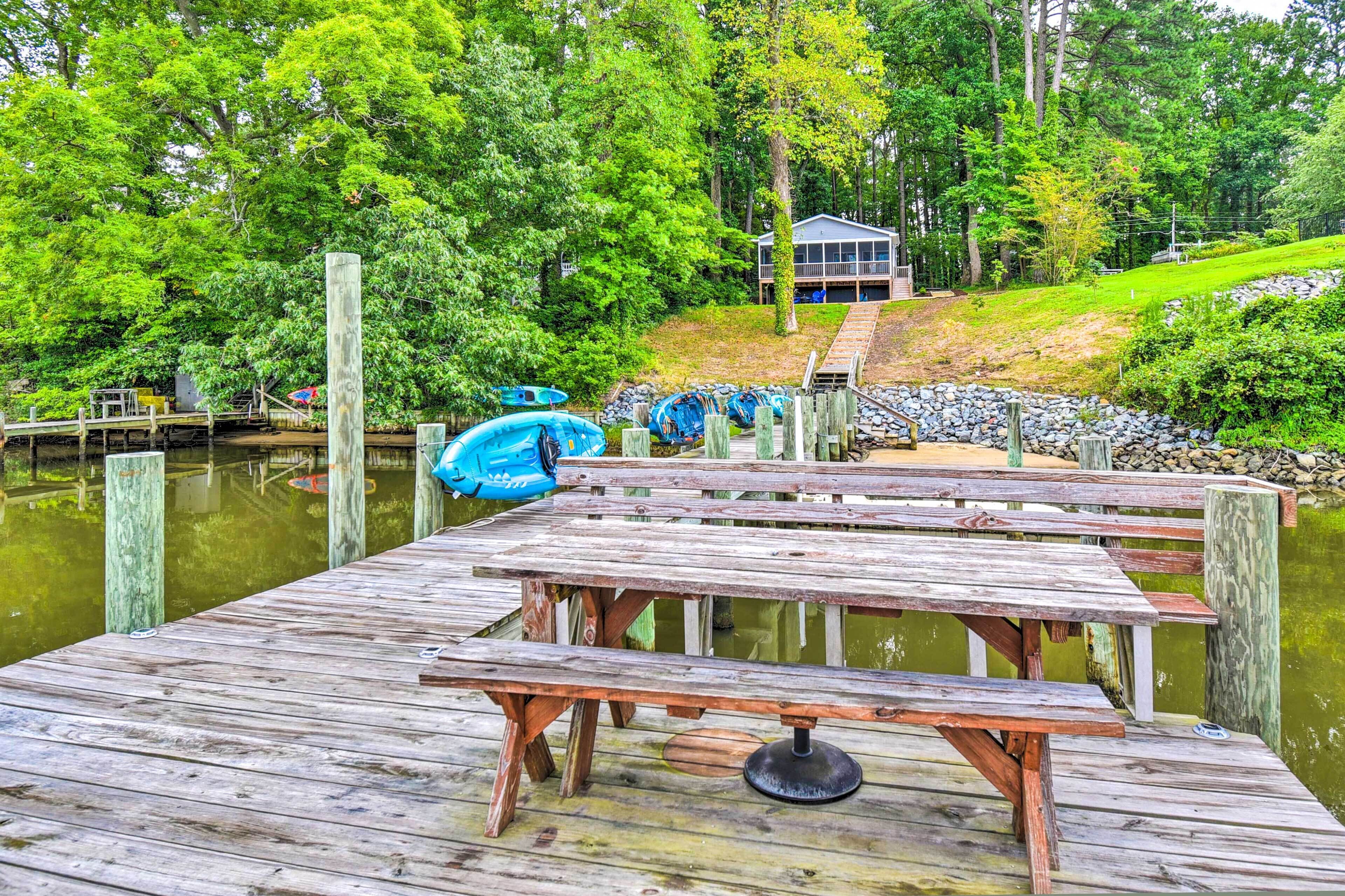 A wooden dock with picnic tables, surrounded by a lake and trees, leading to a house with a porch.