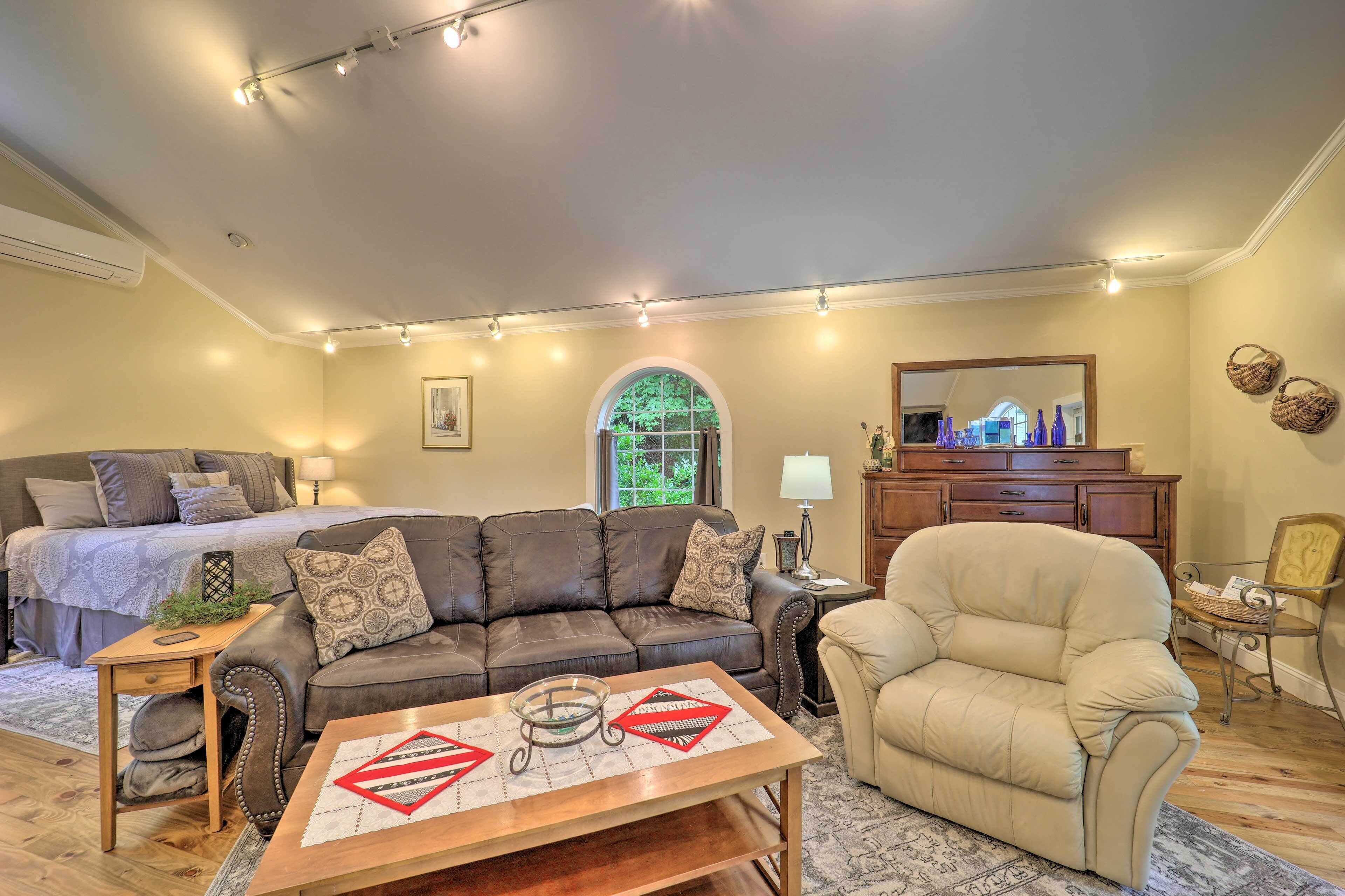 A cozy living room with a sofa, armchair, and a coffee table. There is a bed with a headboard and a dresser with a mirror.