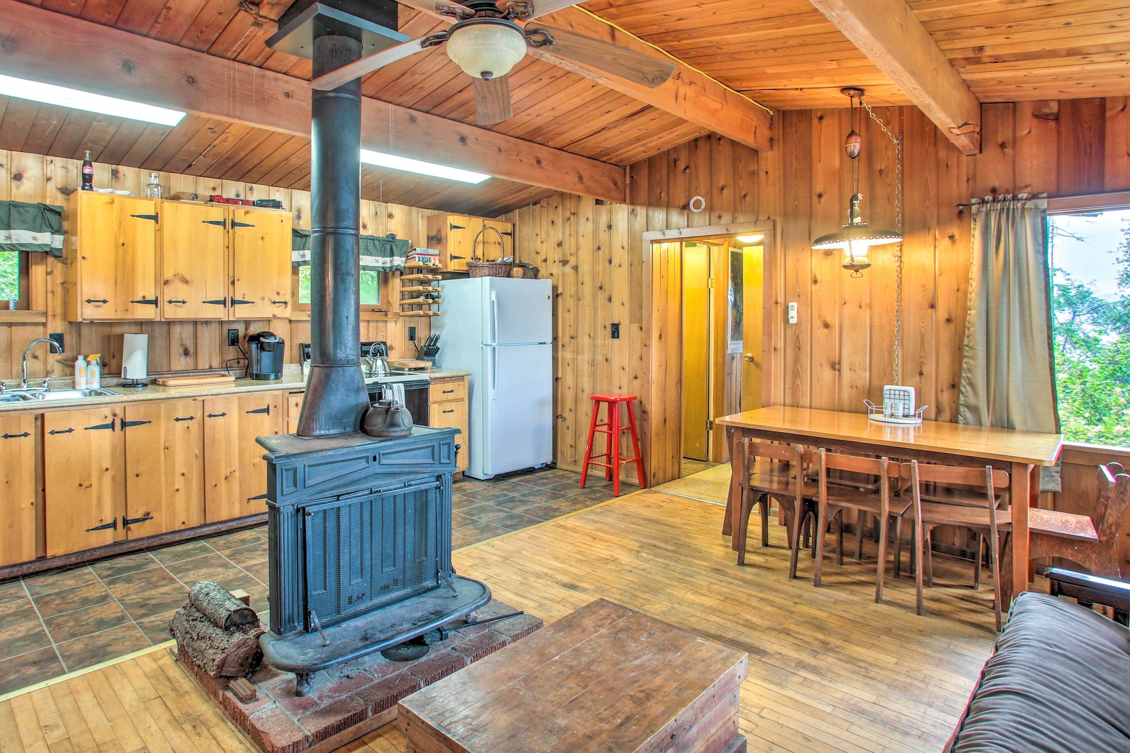 Una baita in legno con cucina, dotata di stufa a legna, zona pranzo con tavolo e sedie, e un'ampia finestra con tende.