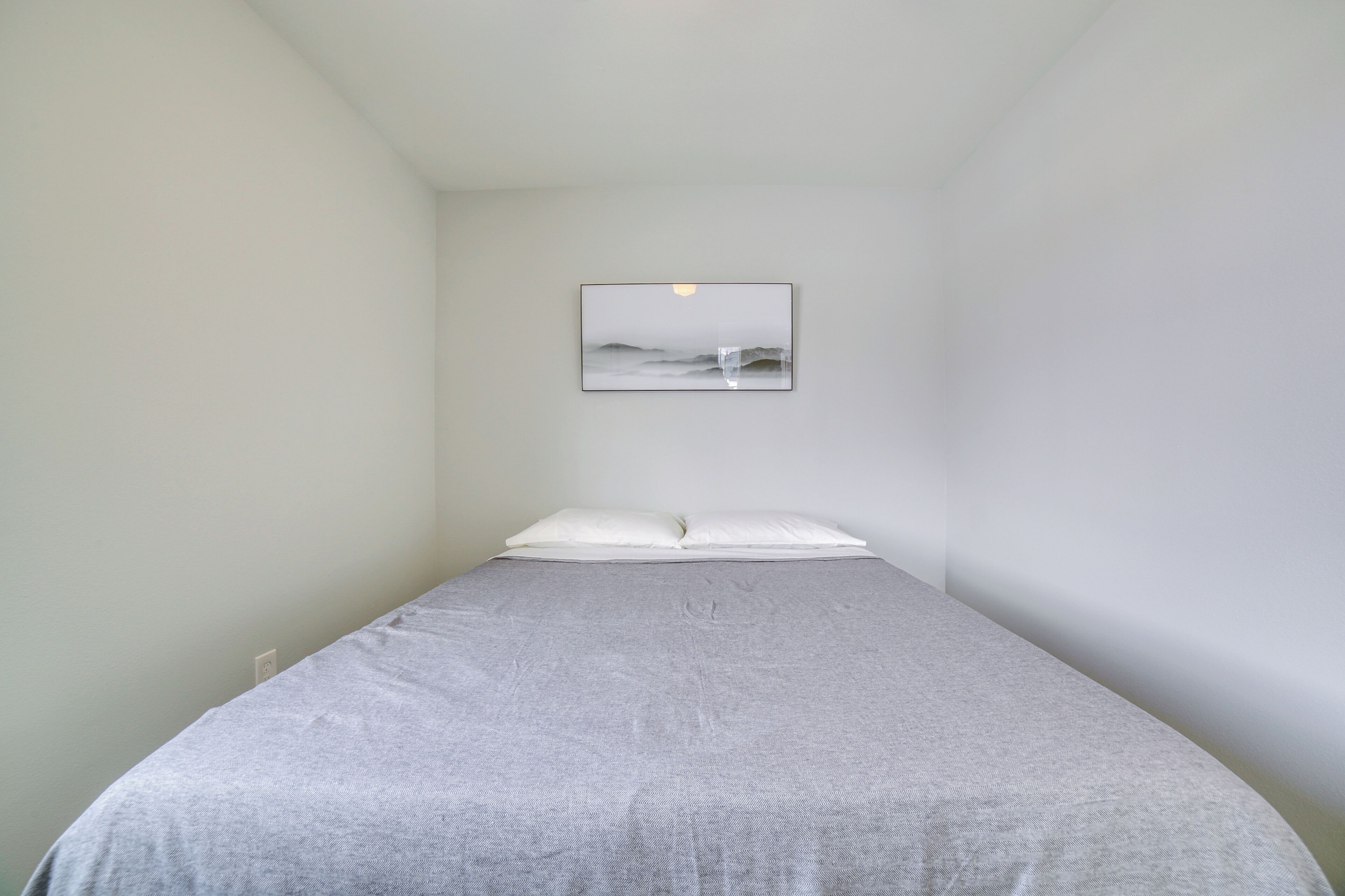 Una habitación pequeña con cama individual, cubrecama gris, almohada blanca y un cuadro en la pared.