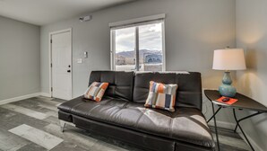 Ein modernes Wohnzimmer mit einer Ledersofa, zwei Zierkissen, einem Beistelltisch mit Lampe und Bergblick durch ein großes Fenster.