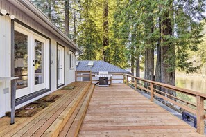 A wooden deck with a grill and a small building, surrounded by tall trees and a body of water. - Riverfront Crescent City Home, 8 Mi to Beach! (Crescent City)