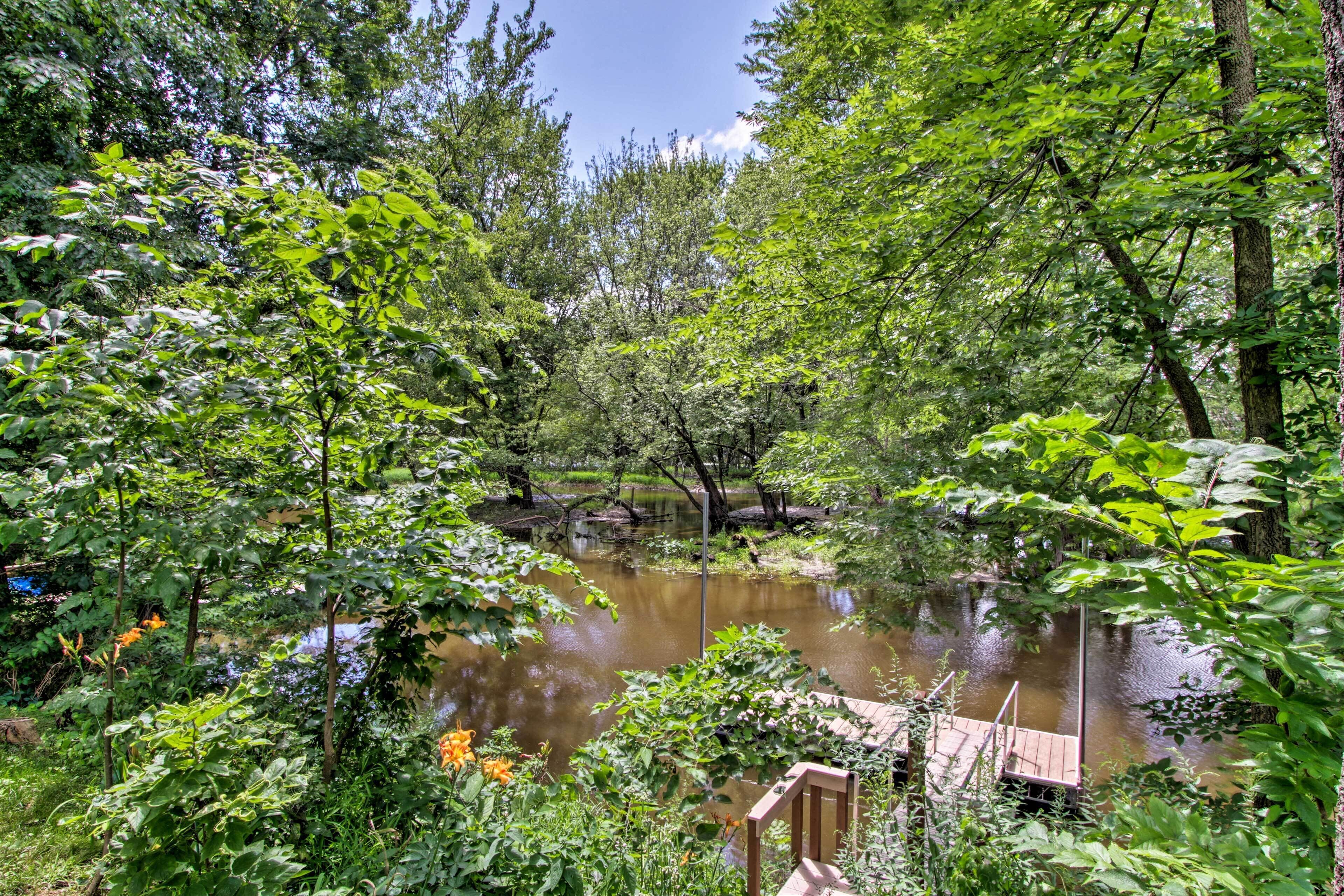 A serene riverside setting with a wooden dock extending into the water, surrounded by lush greenery and trees.