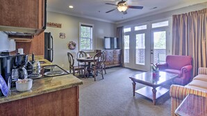 A spacious living area with a dining table, chairs, a coffee table, and a red armchair. There is a kitchen area with a stove, sink, and cabinets. A ceiling fan and a TV are visible in the background. - Resort-style Condo/suite on Golf Course w/ Pool! (Spring Lake)