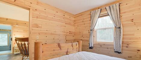 Una habitación con paneles de madera, una cama, un sillón mecededor y una ventana con cortinas estampadas.