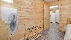 Une pièce aux murs lambrés, équipée d’un ventilateur sur pied, d’un porte-vêtements et d’un lit.