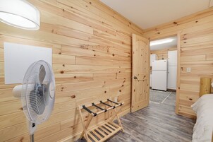 Una habitación con paneles de madera, un ventilador de pie, un perchero y una cama.