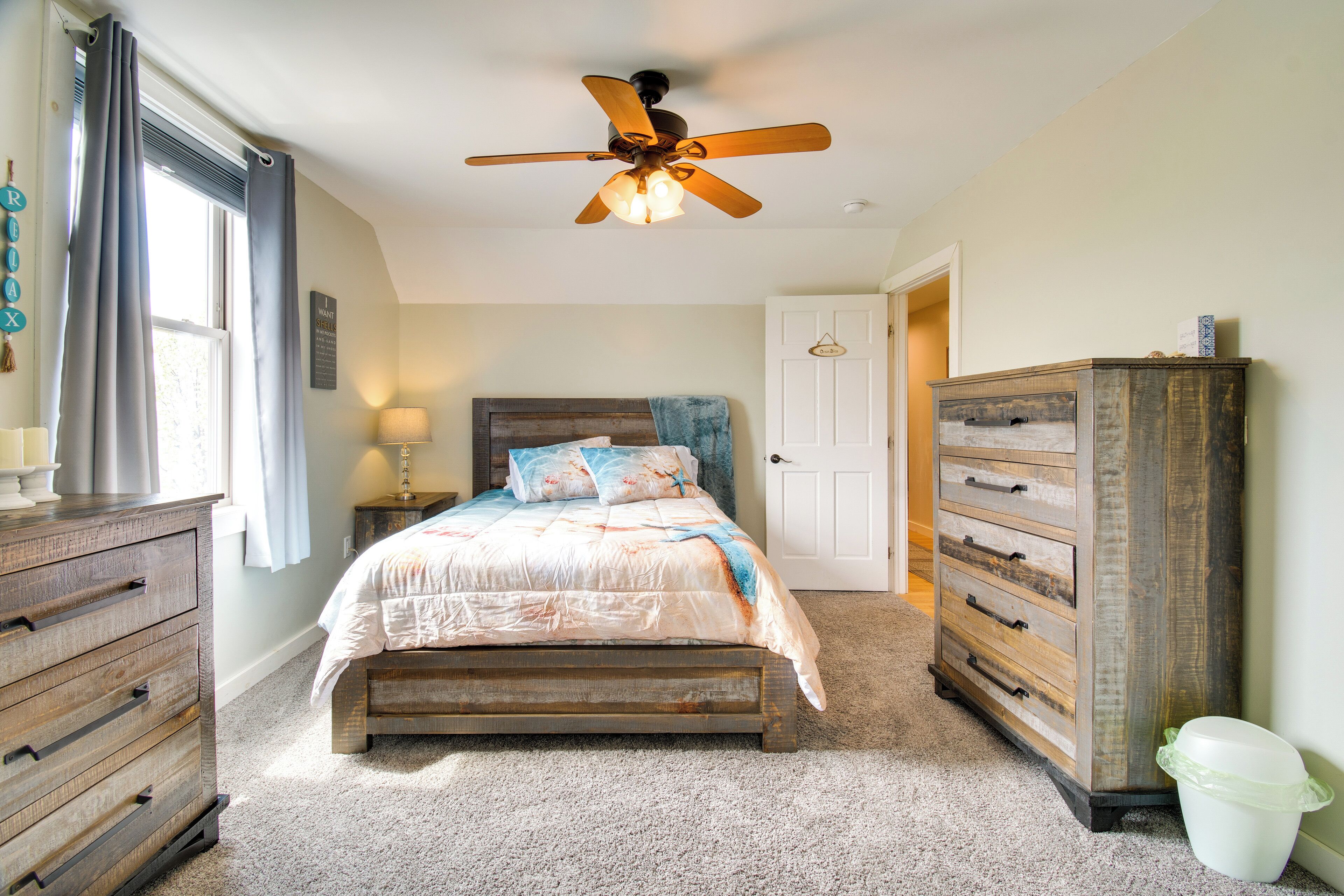 A bedroom with a bed, a wooden dresser, a nightstand, a ceiling fan, and a window with curtains.