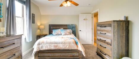 A bedroom with a bed, a wooden dresser, a nightstand, a ceiling fan, and a window with curtains.