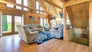 A log cabin interior with a wooden ceiling, large windows, a living room with sofas and armchairs, and a view of the outdoors. - Rural New York Vacation Rental w/ Deck (Romulus)
