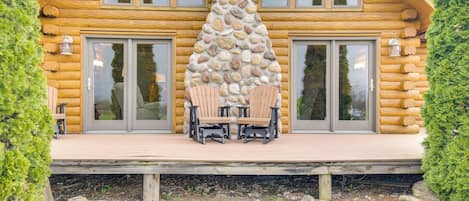 A log cabin with a stone chimney, large windows, and a deck with chairs.