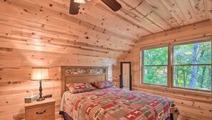 Un dormitorio de cabaña de madera con una cama, una mesita de noche, una lámpara y un ventanal con vista al bosque.