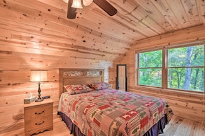 Un dormitorio de cabaña de madera con una cama, una mesita de noche, una lámpara y un ventanal con vista al bosque.